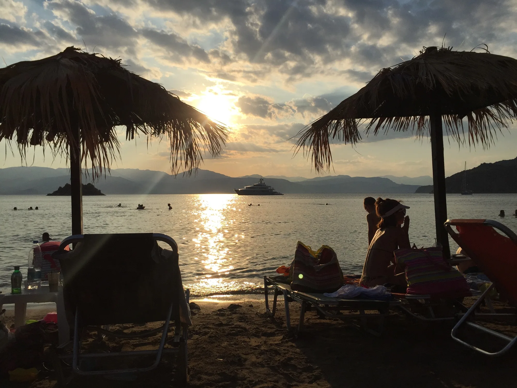 6 παραλίες με φυσική σκιά στην Πελοπόννησο Ηλιοβασίλεμα στην παραλία Καραθώνα με ψάθινες ομπρέλες, λουόμενους και σκάφος στον Αργολικό Κόλπο