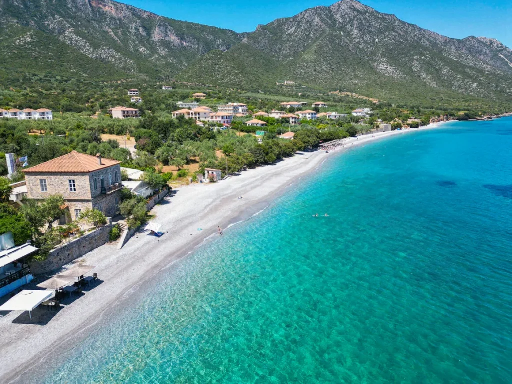 6 παραλίες με φυσική σκιά στην Πελοπόννησο Αεροφωτογραφία της βοτσαλωτής παραλίας των Πουλίθρων με γαλαζοπράσινα νερά, πέτρινα σπίτια και καταπράσινες πλαγιές