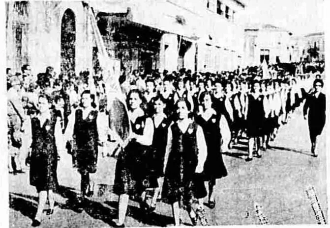 28η Οκτωβρίου στην Πάτρα: Όταν το ’40 καθόταν απέναντι από την εξέδρα Μαθήτριες με σκούρες στολές και λευκούς γιακάδες παρελαύνουν με τη σημαία, ενώ θεατές παρακολουθούν από το πεζοδρόμιο