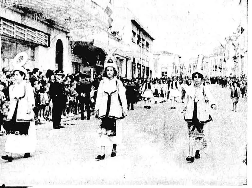 28η Οκτωβρίου στην Πάτρα: Όταν το ’40 καθόταν απέναντι από την εξέδρα Γυναίκες με παραδοσιακές φορεσιές προπορεύονται σε τμήμα της παρέλασης, με θεατές και παλιά κτίρια στο βάθος.