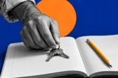 Ασπρόμαυρο ενήλικο χέρι ακουμπά κλειδιά σε ανοιχτό τετράδιο με μολύβι, σε μπλε και πορτοκαλί φόντο.